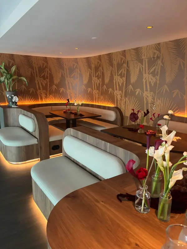 Elegant restaurant interior with curved white leather booths, bamboo wallpaper, warm lighting, and fresh floral centerpieces.